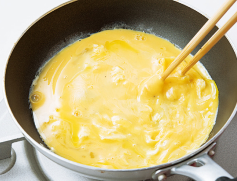 等蛋液從邊緣開始凝結後，左手前後搖晃平底鍋，右手用長筷子在鍋裡攪動。